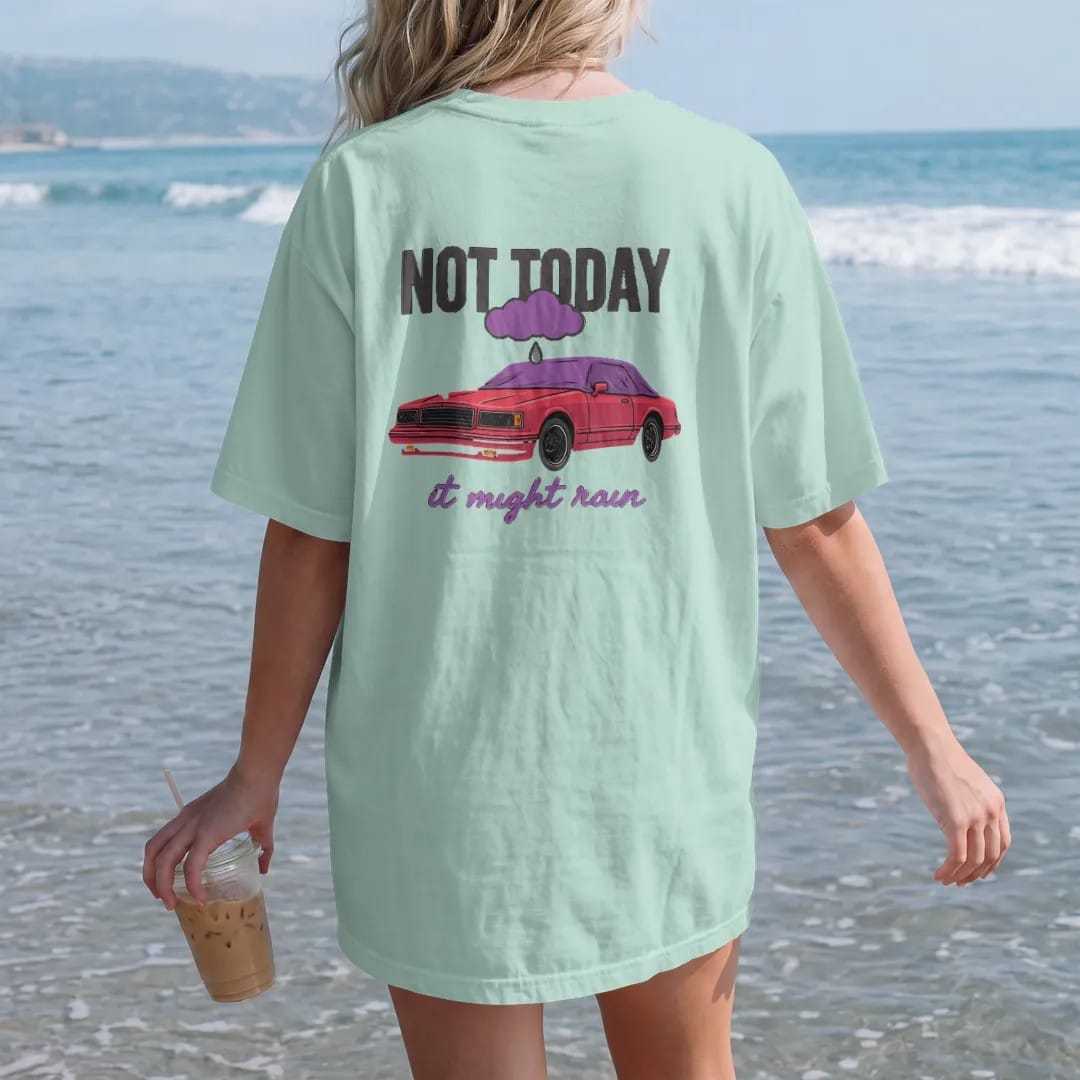 Woman on the beach in a Chalky Mint tee, back view: a boxy sedan under a car cover in a carport beneath a little rain cloud, with Not Today over It Might Rain, in deep red, purple and gunmetal.