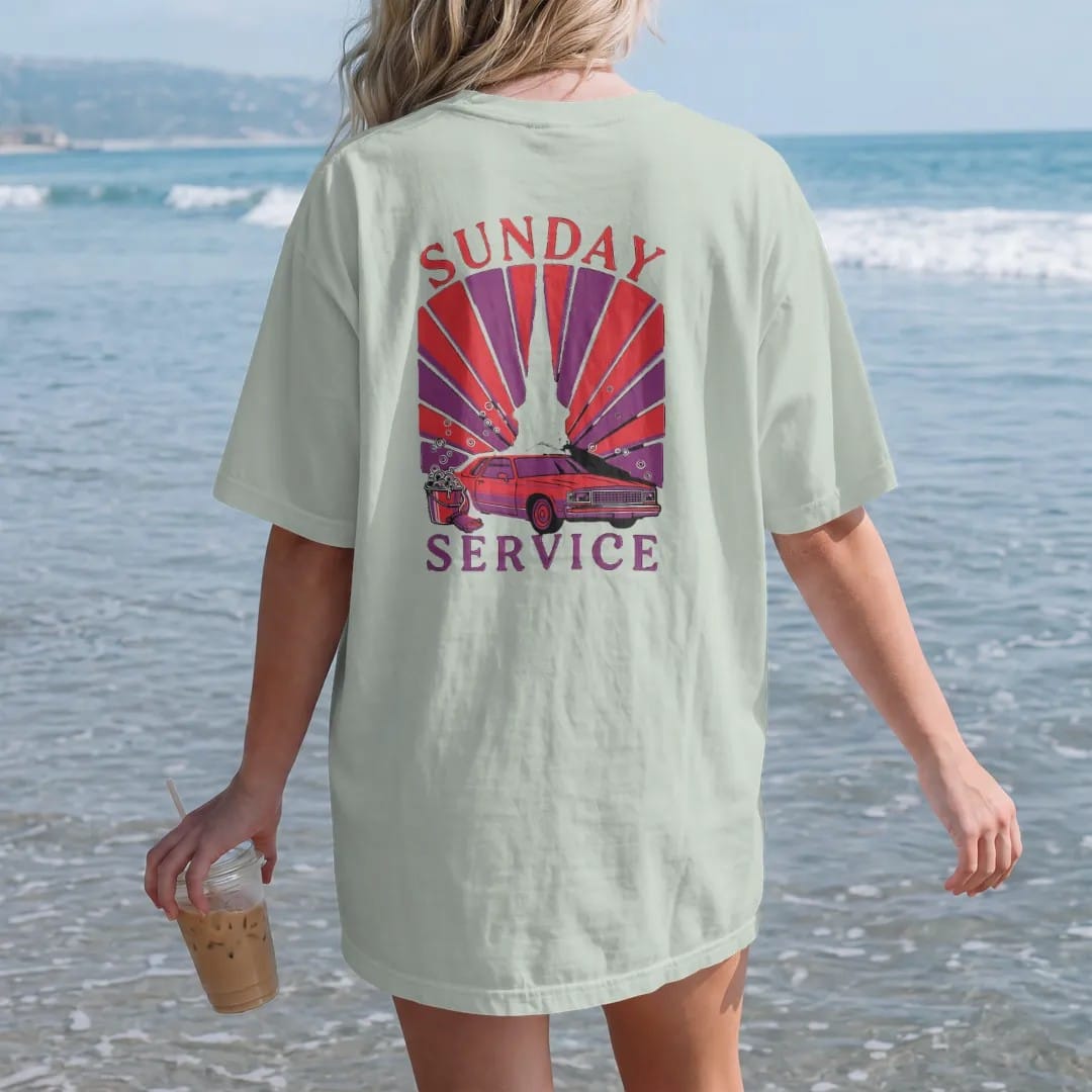 Woman on the beach in a Bay tee, back view: a church steeple behind a boxy sedan on chrome rims being washed with a soap bucket and mitt, under Sunday and over Service, in deep red, purple and gunmetal.