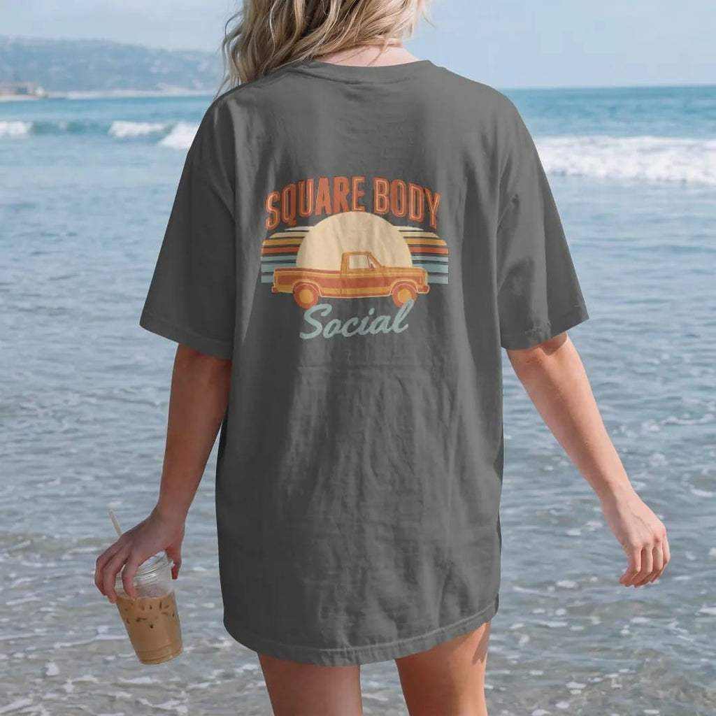 Woman on the beach in a Pepper tee, back view: a two-tone square-body pickup in side profile against a faded 70s rainbow sunset, under Square Body and over Social in cream, gold, orange and teal.