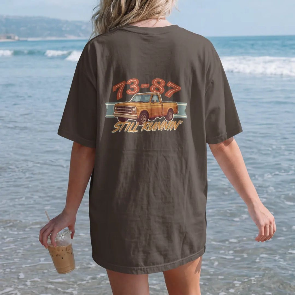 Woman on the beach in a Espresso tee, back view: a square-body pickup in front three-quarter view with 73-87 in big retro numerals above and Still Runnin' in brush script below.