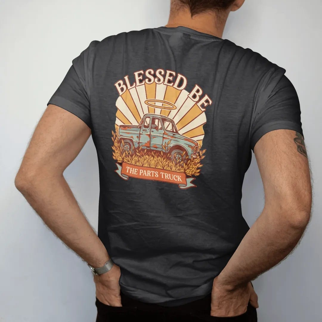Man wearing a Black tee, seen from the back: a rusty square-body pickup in tall weeds with a glowing halo and light rays, under Blessed Be and over a banner reading The Parts Truck.