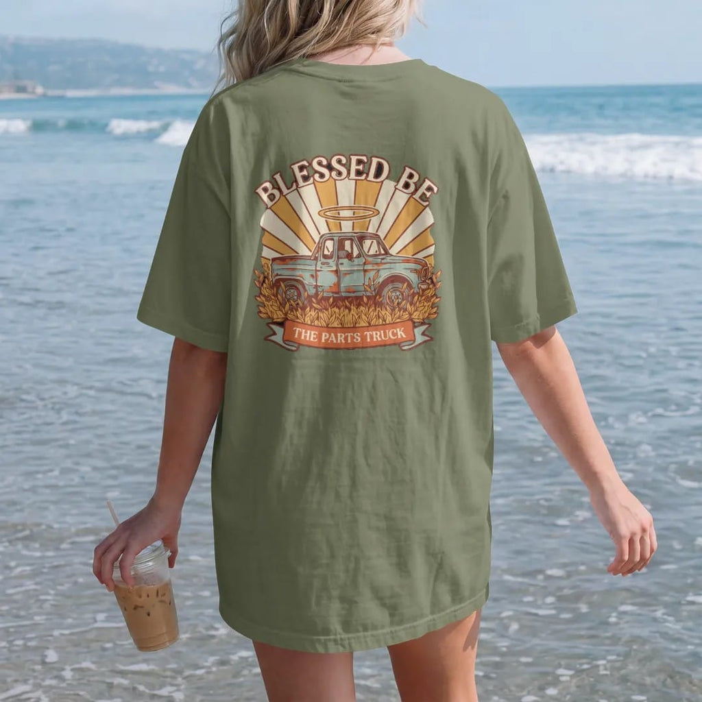 Woman on the beach in a Moss tee, back view: a rusty square-body pickup in tall weeds with a glowing halo and light rays, under Blessed Be and over a banner reading The Parts Truck.