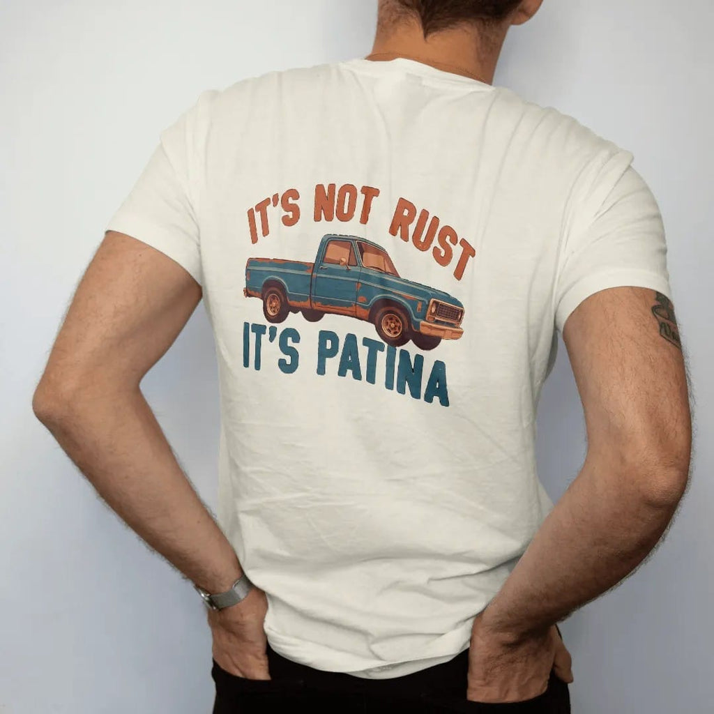 Man wearing a Ivory tee, seen from the back: a weathered teal square-body pickup with rust patches, It's Not Rust above and It's Patina below in bold block letters.