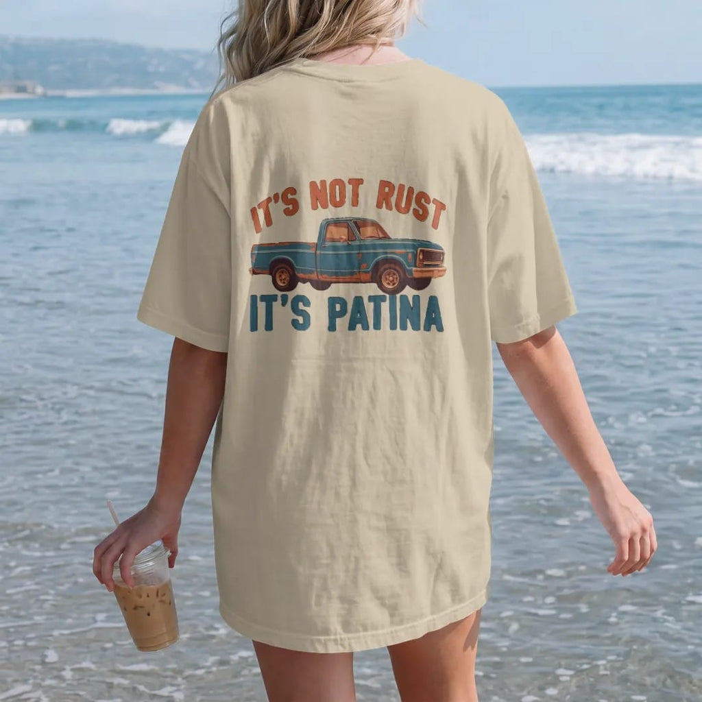 Woman on the beach in a Sandstone tee, back view: a weathered teal square-body pickup with rust patches, It's Not Rust above and It's Patina below in bold block letters.
