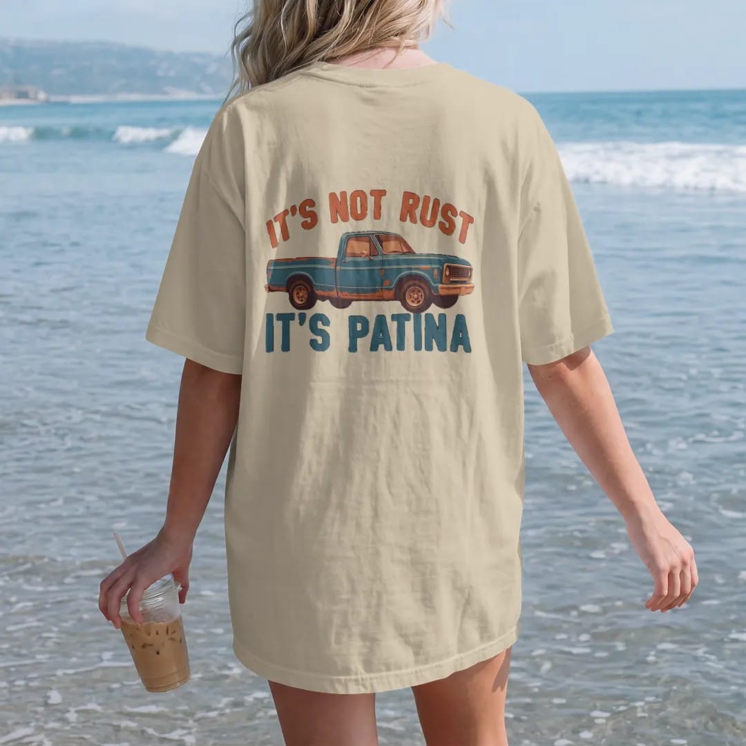 Woman on the beach in a Sandstone tee, back view: a weathered teal square-body pickup with rust patches, It's Not Rust above and It's Patina below in bold block letters.