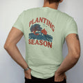 Man wearing a Light Green tee, seen from the back: a square-body pickup with seed sacks in the bed beside plowed rows, a dogwood and a red barn, under Planting and over Season, in rust, teal and navy.