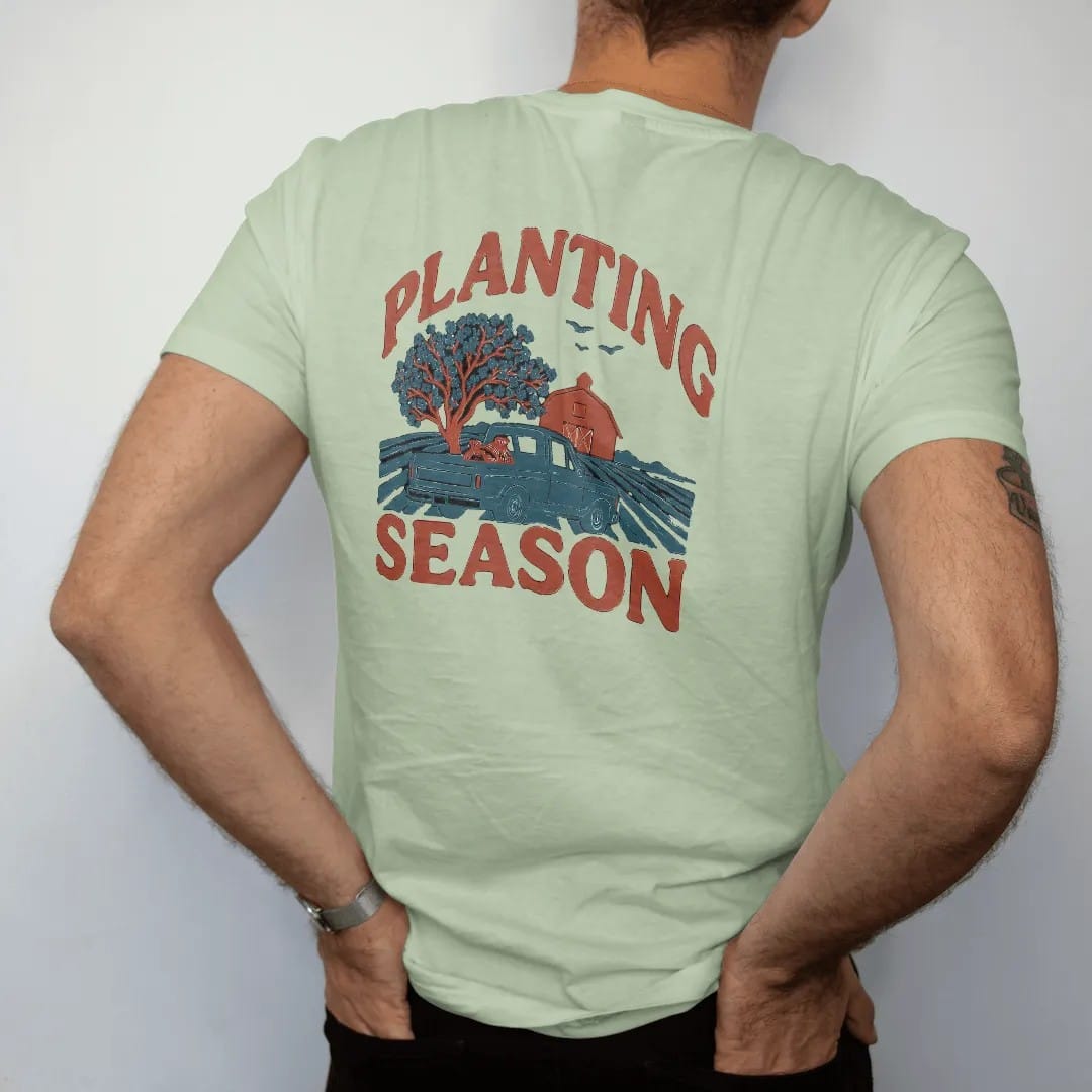 Man wearing a Light Green tee, seen from the back: a square-body pickup with seed sacks in the bed beside plowed rows, a dogwood and a red barn, under Planting and over Season, in rust, teal and navy.