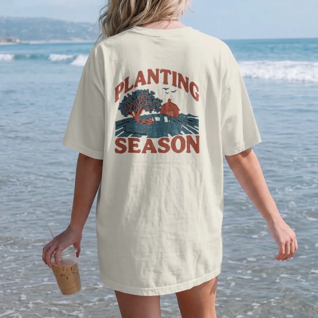 Woman on the beach in a Ivory tee, back view: a square-body pickup with seed sacks in the bed beside plowed rows, a dogwood and a red barn, under Planting and over Season, in rust, teal and navy.