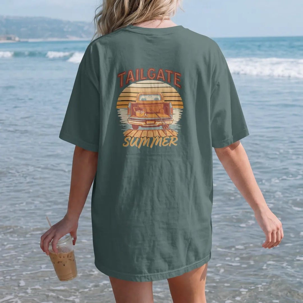 Woman on the beach in a Blue Spruce tee, back view: a square-body pickup at a lake dock with the tailgate down, cooler and fishing poles, against a striped sunset, under Tailgate and over Summer.