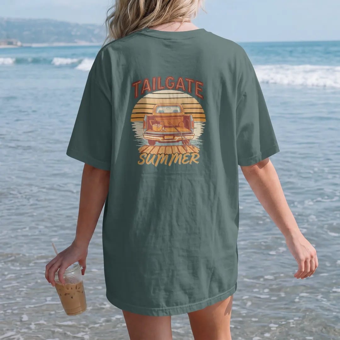 Woman on the beach in a Blue Spruce tee, back view: a square-body pickup at a lake dock with the tailgate down, cooler and fishing poles, against a striped sunset, under Tailgate and over Summer.