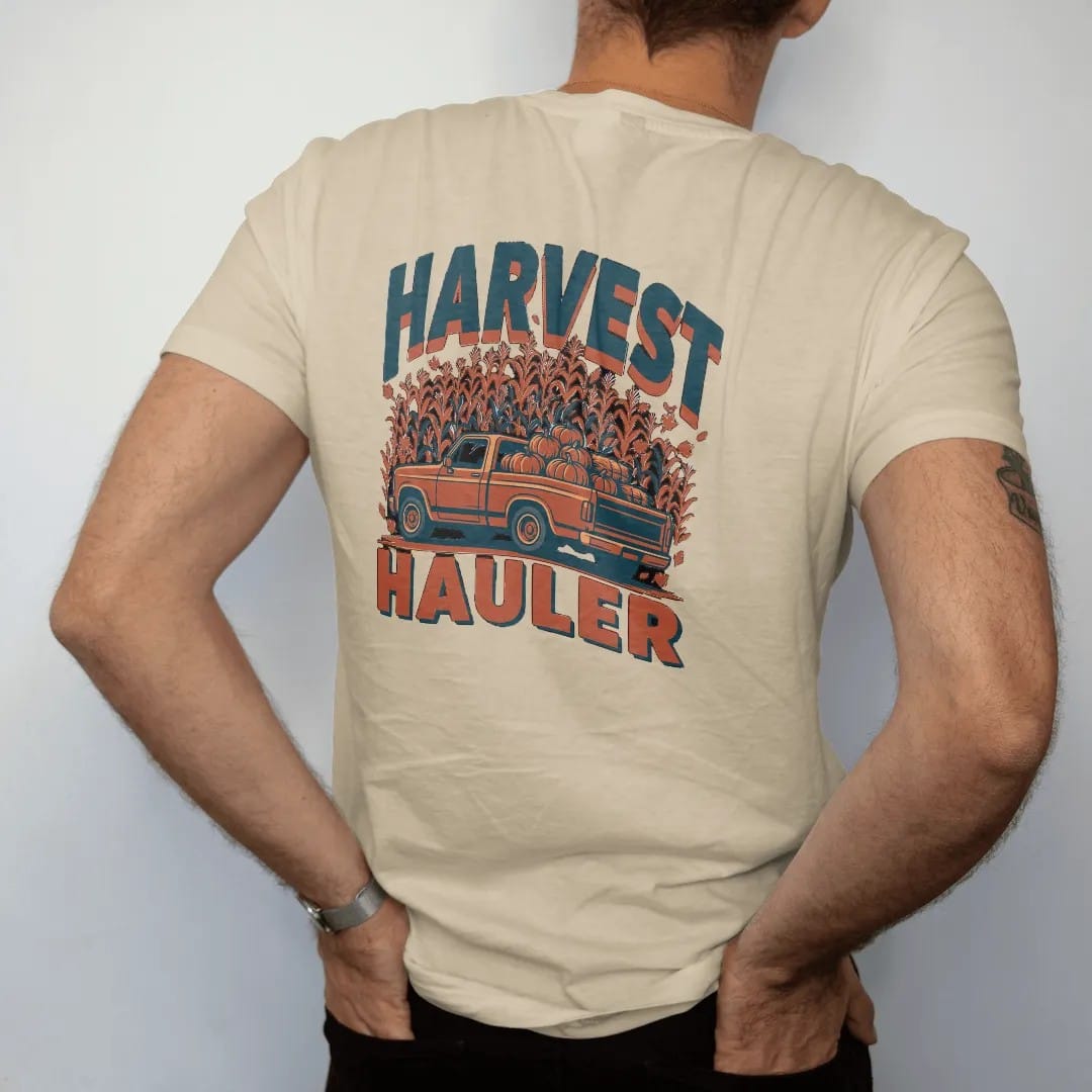 Man wearing a Sandstone tee, seen from the back: a square-body pickup piled with pumpkins and hay bales among falling leaves and corn, under Harvest and over Hauler, in rust, teal and navy.