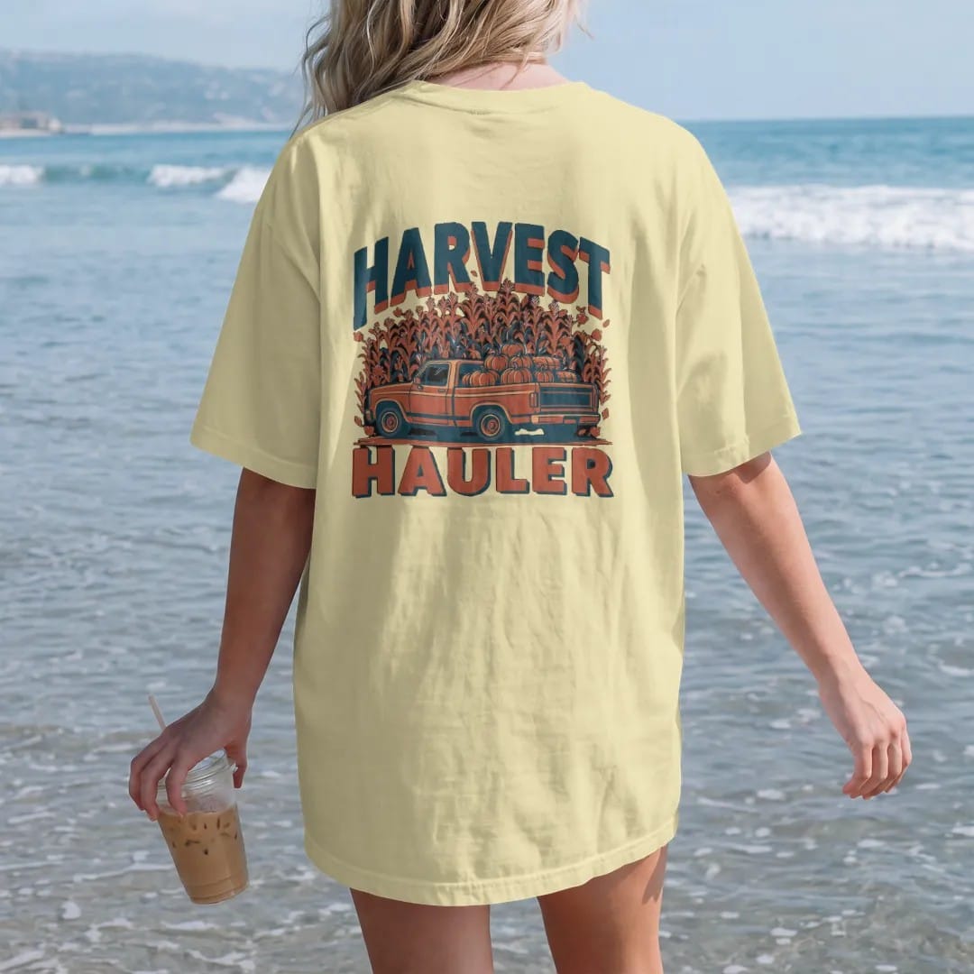 Woman on the beach in a Butter tee, back view: a square-body pickup piled with pumpkins and hay bales among falling leaves and corn, under Harvest and over Hauler, in rust, teal and navy.