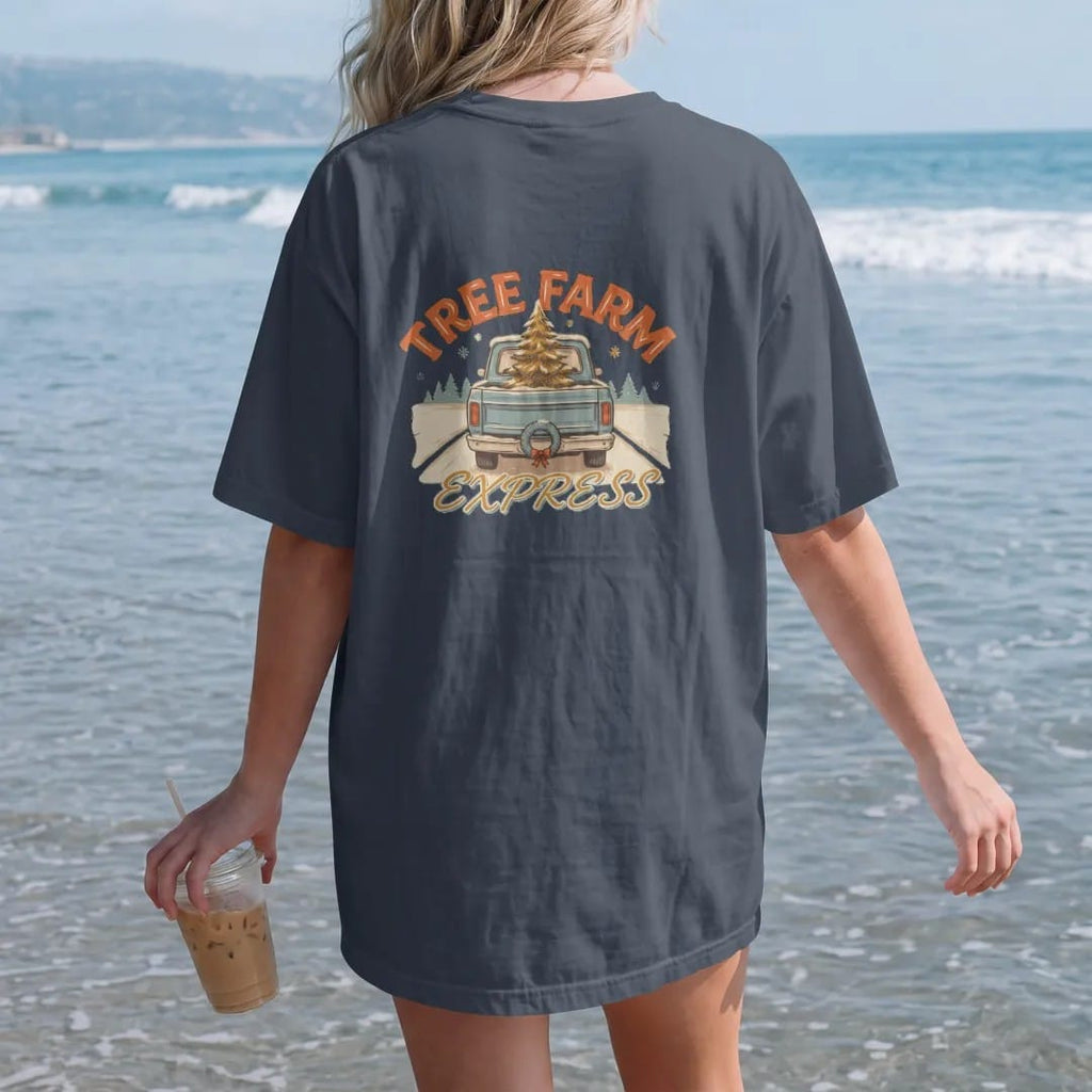 Woman on the beach in a Midnight tee, back view: a square-body pickup on a snowy road with a Christmas tree in the bed and a wreath, under Tree Farm and over Express in retro script.