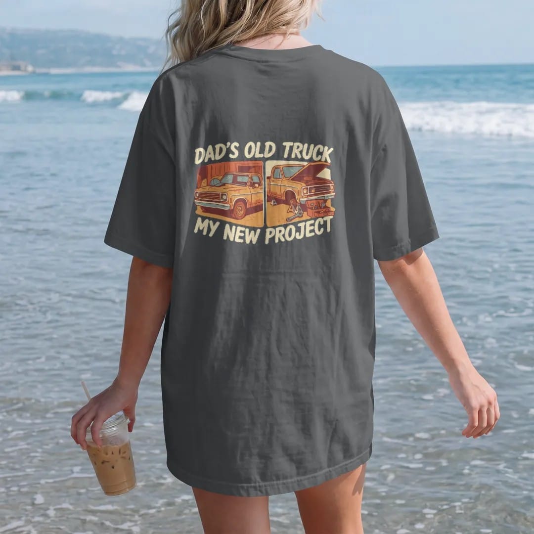 Woman on the beach in a Graphite tee, back view: a square-body pickup shown parked in a barn and then on jack stands with a toolbox, under Dad's Old Truck and over My New Project.