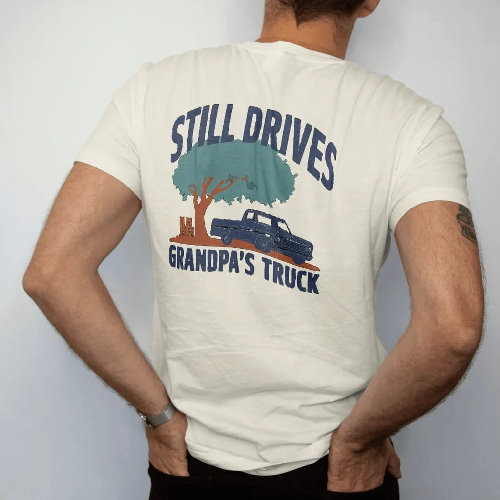 Man wearing a Ivory tee, seen from the back: a navy square-body pickup parked under a big shade tree by a porch, with Still Drives above and Grandpa's Truck below.