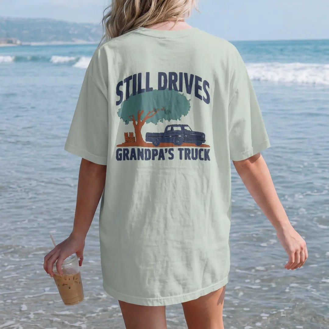 Woman on the beach in a Bay tee, back view: a navy square-body pickup parked under a big shade tree by a porch, with Still Drives above and Grandpa's Truck below.