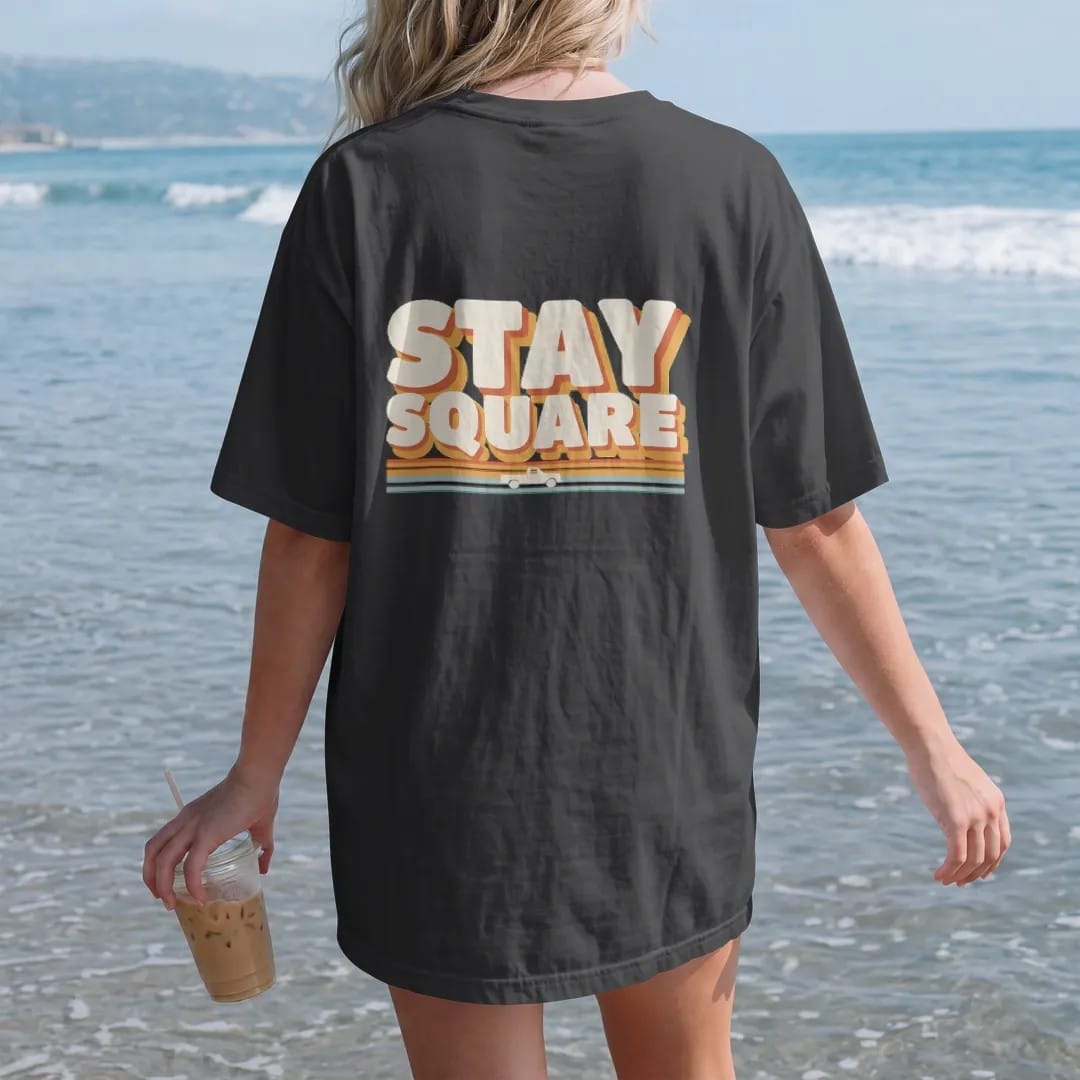 Woman on the beach in a Black tee, back view: a tiny square-body pickup riding 70s sunset stripes under Stay Square in giant cream block letters with an orange and gold drop shadow.