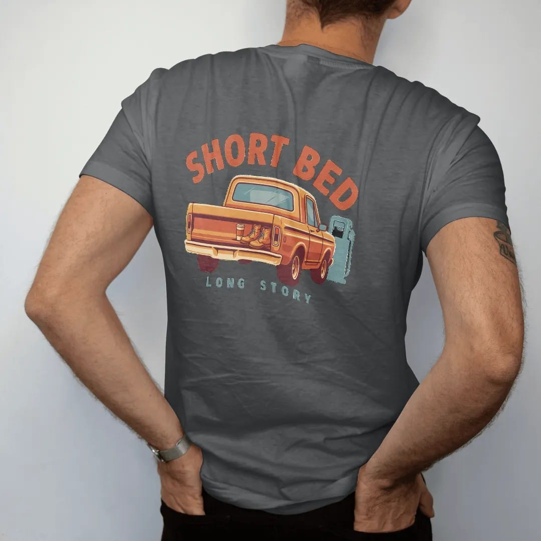 Man wearing a Graphite tee, seen from the back: a short-bed square-body pickup at a gas station at dusk with boots and coffee on the tailgate, under Short Bed and over Long Story.