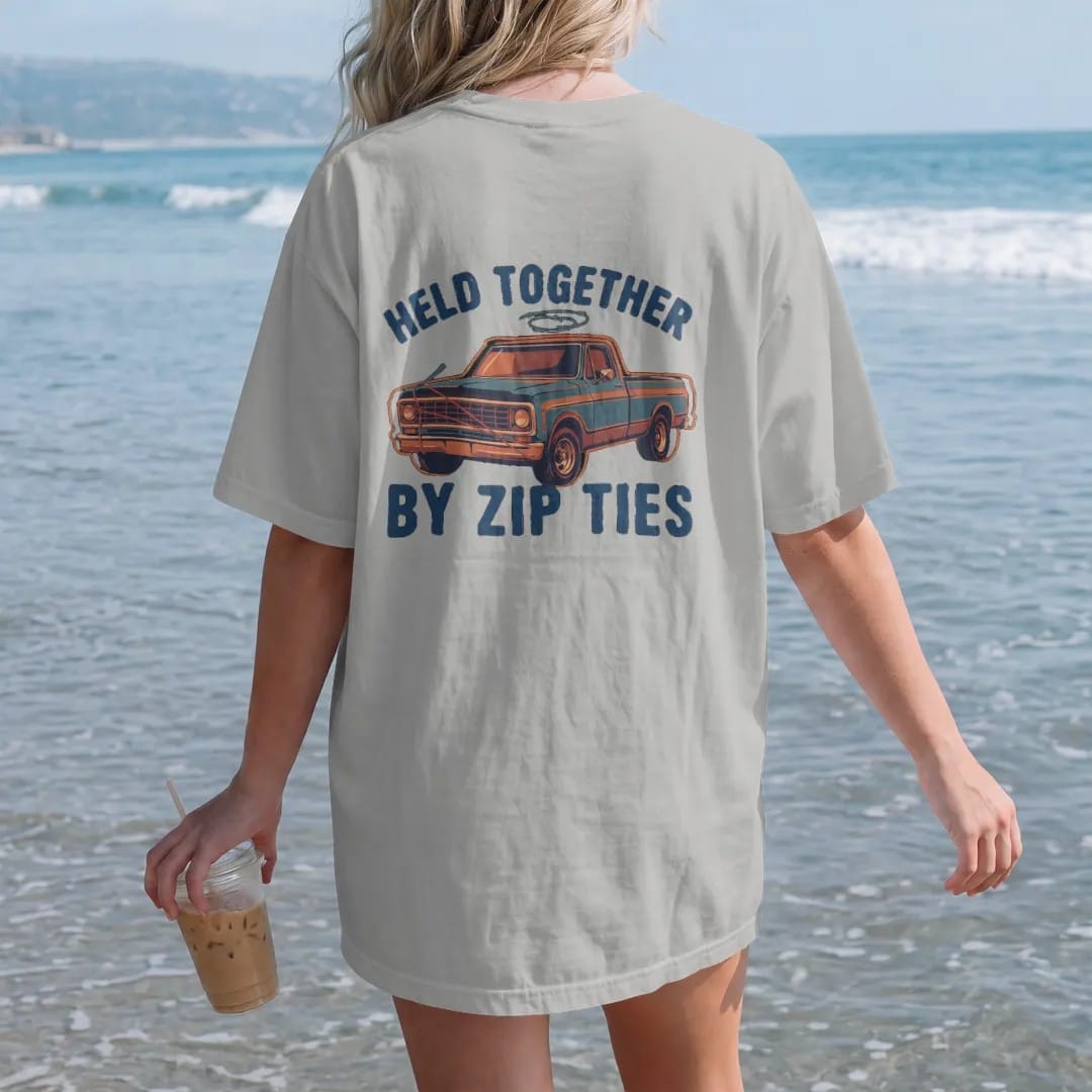 Woman on the beach in a Granite tee, back view: a square-body pickup with a mismatched fender, bungee-strapped hood and zip ties everywhere, under Held Together and over By Zip Ties, in rust, teal and navy.