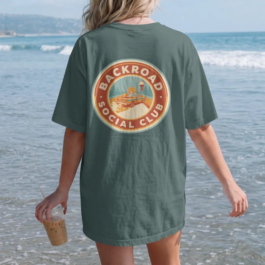 Woman on the beach in a Blue Spruce tee, back view: a round road-trip badge of a square-body pickup on a dirt road past pines and a water tower, with Backroad arched over the top and Social Club around the bottom.