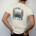 Man wearing a Ivory tee, seen from the back: a dusty square-body pickup covered in hay with a cat on the hood inside open barn doors, under Found It and over In A Barn, in rust, teal and navy.