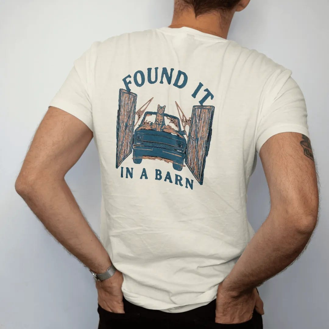 Man wearing a Ivory tee, seen from the back: a dusty square-body pickup covered in hay with a cat on the hood inside open barn doors, under Found It and over In A Barn, in rust, teal and navy.