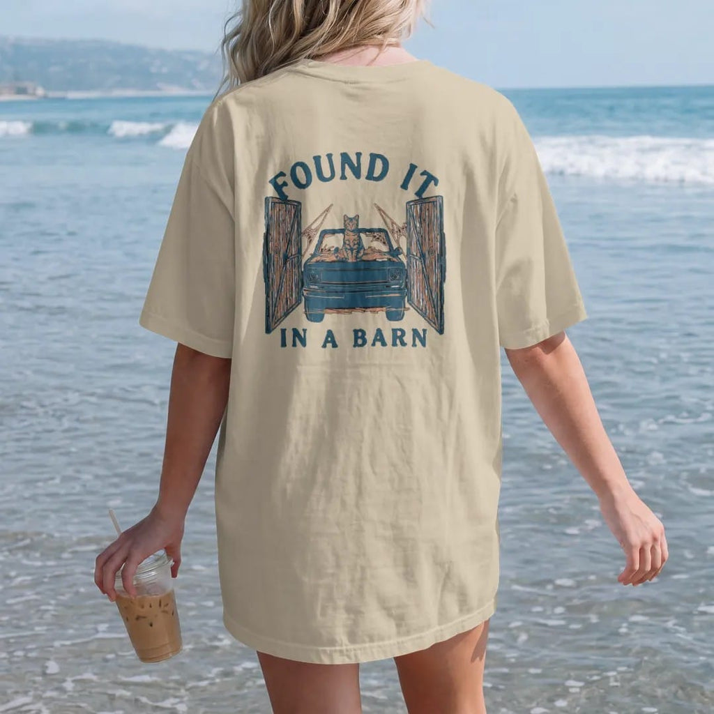 Woman on the beach in a Sandstone tee, back view: a dusty square-body pickup covered in hay with a cat on the hood inside open barn doors, under Found It and over In A Barn, in rust, teal and navy.