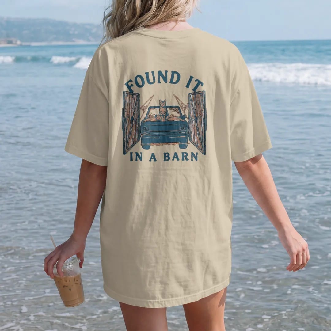 Woman on the beach in a Sandstone tee, back view: a dusty square-body pickup covered in hay with a cat on the hood inside open barn doors, under Found It and over In A Barn, in rust, teal and navy.