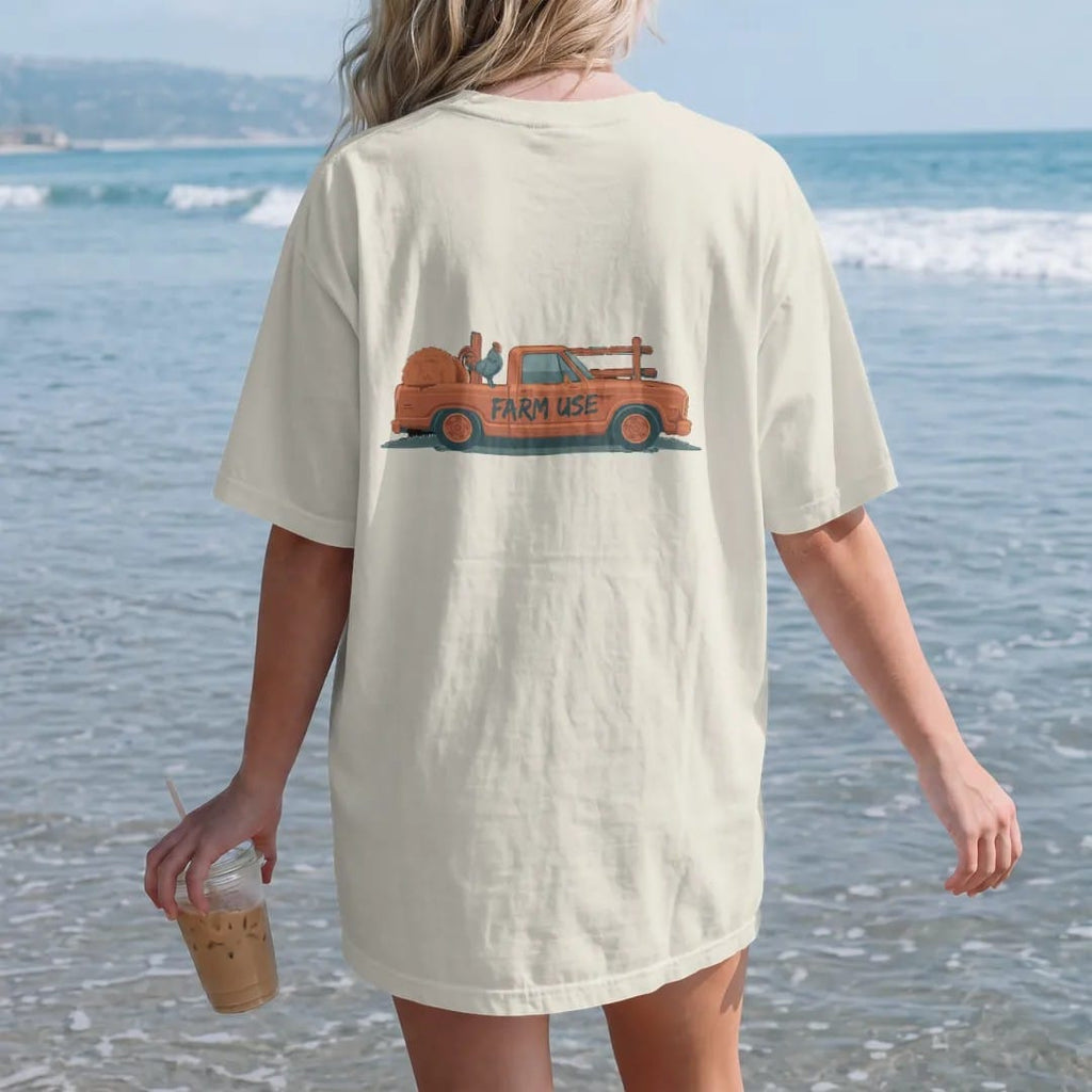 Woman on the beach in a Ivory tee, back view: a dented farm square-body pickup with Farm Use hand-painted on the door, a hay bale and a rooster in the bed, in rust, teal and navy.