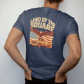 Man wearing a Navy tee, seen from the back: an eagle soaring over a faded American flag and a square-body pickup, with Land Of The Square arched above, in vintage rust, gold and cream.