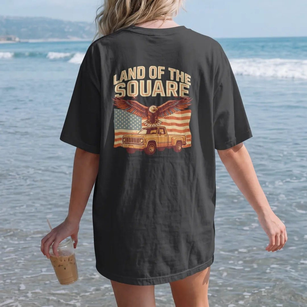 Woman on the beach in a Black tee, back view: an eagle soaring over a faded American flag and a square-body pickup, with Land Of The Square arched above, in vintage rust, gold and cream.