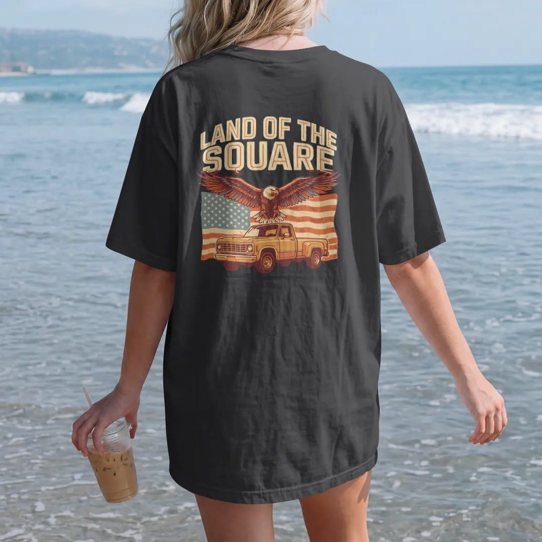 Woman on the beach in a Black tee, back view: an eagle soaring over a faded American flag and a square-body pickup, with Land Of The Square arched above, in vintage rust, gold and cream.