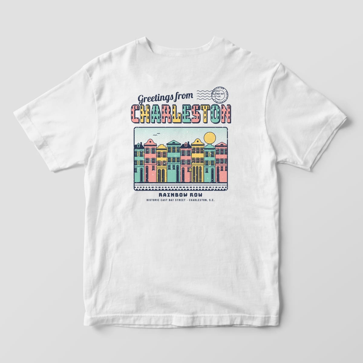 Back of a White Comfort Colors 1717 tee printed with Greetings from Charleston in pastel block letters above a row of pink, yellow and mint row houses.