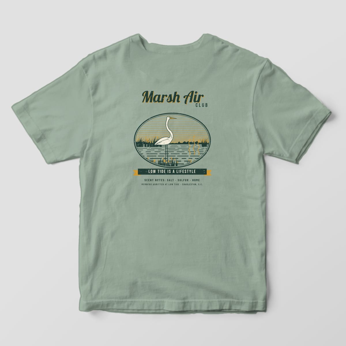 Back of a Bay Comfort Colors 1717 tee with a dark green, cream and gold club badge: a great egret in a marsh oval, aroma lines over the water, and Low Tide Is a Lifestyle.