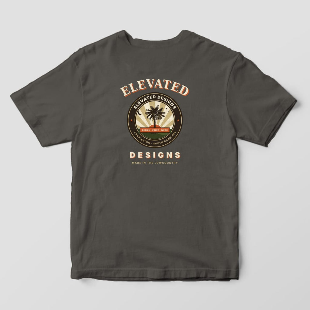 Back of a Pepper Comfort Colors 1717 tee with ELEVATED arched over a round palmetto crest with a sunburst, crescent moon and DESIGN · PRINT · WEAR banner, then DESIGNS and MADE IN THE LOWCOUNTRY below.
