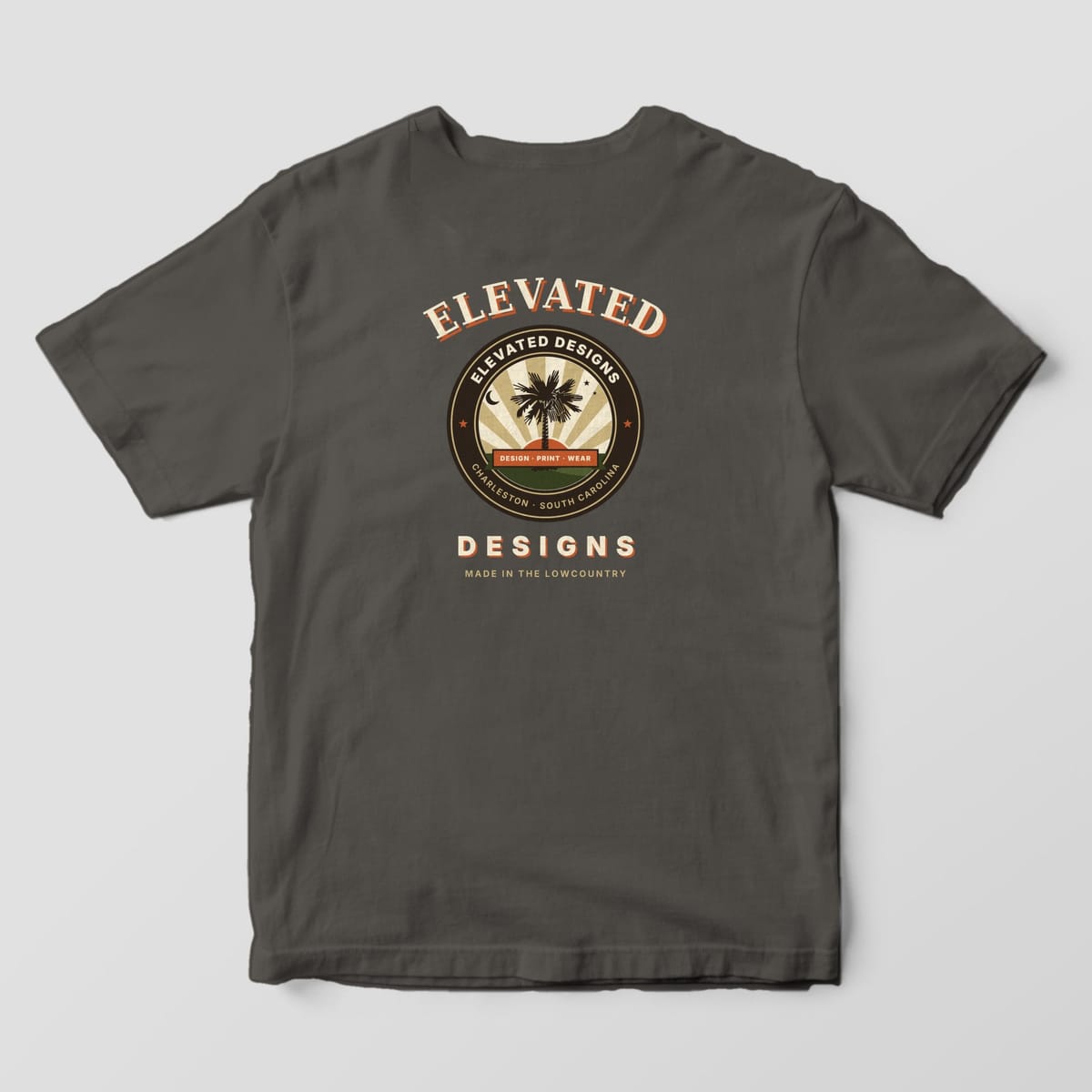 Back of a Pepper Comfort Colors 1717 tee with ELEVATED arched over a round palmetto crest with a sunburst, crescent moon and DESIGN · PRINT · WEAR banner, then DESIGNS and MADE IN THE LOWCOUNTRY below.