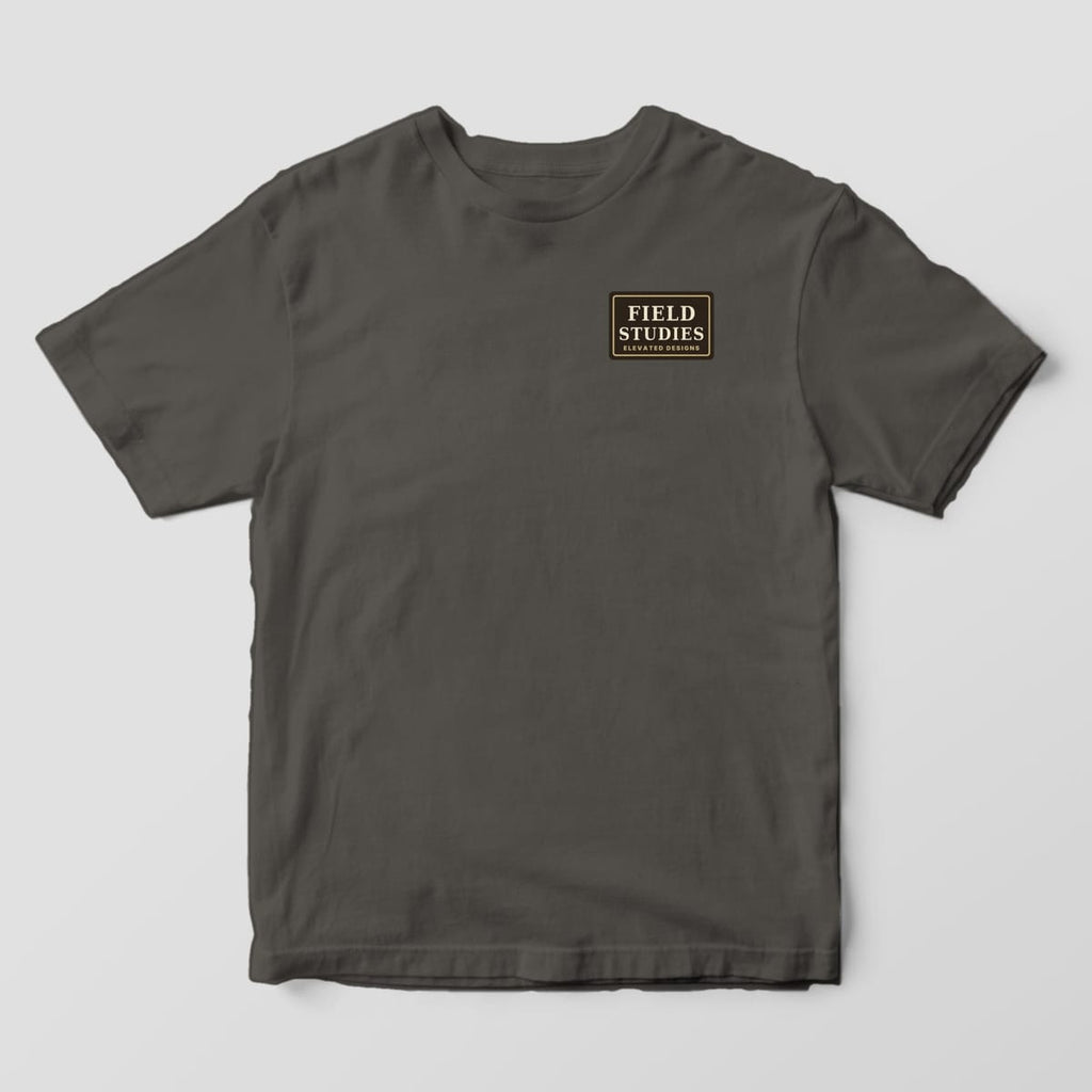 Front of a Pepper Comfort Colors 1717 tee with a small dark brown left-chest label with a tan border reading FIELD STUDIES, ELEVATED DESIGNS.