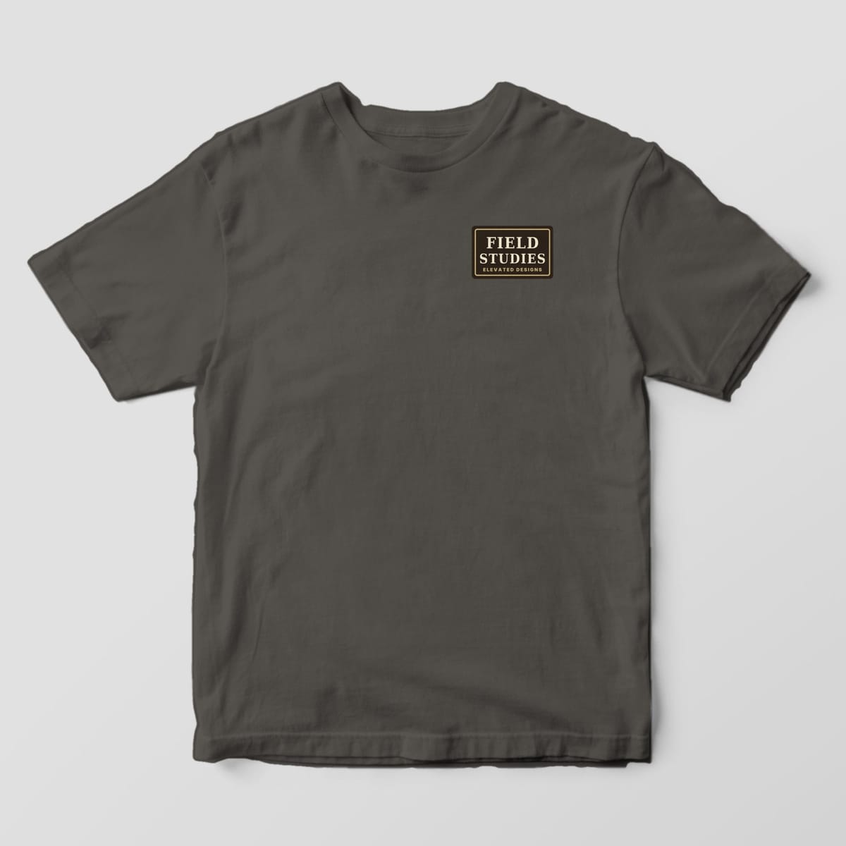 Front of a Pepper Comfort Colors 1717 tee with a small dark brown left-chest label with a tan border reading FIELD STUDIES, ELEVATED DESIGNS.
