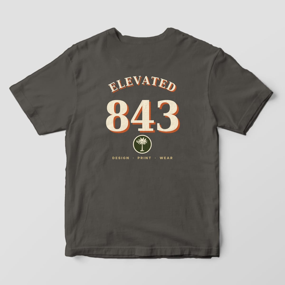 Back of a Pepper Comfort Colors 1717 tee with ELEVATED arched over oversized cream-and-rust 843 numbers, a small olive palmetto and crescent patch, and DESIGN · PRINT · WEAR.