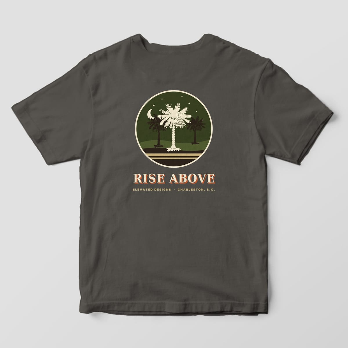 Back of a Pepper Comfort Colors 1717 tee with a round olive night scene of three palmetto trees under a crescent moon and stars, with RISE ABOVE and ELEVATED DESIGNS · CHARLESTON, S.C. below.