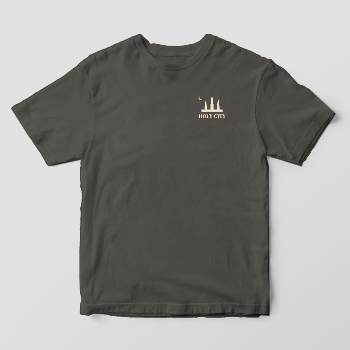 Front of a Pepper Comfort Colors 1717 tee with a small left-chest cream skyline of three steeples and a gold crescent moon above HOLY CITY.