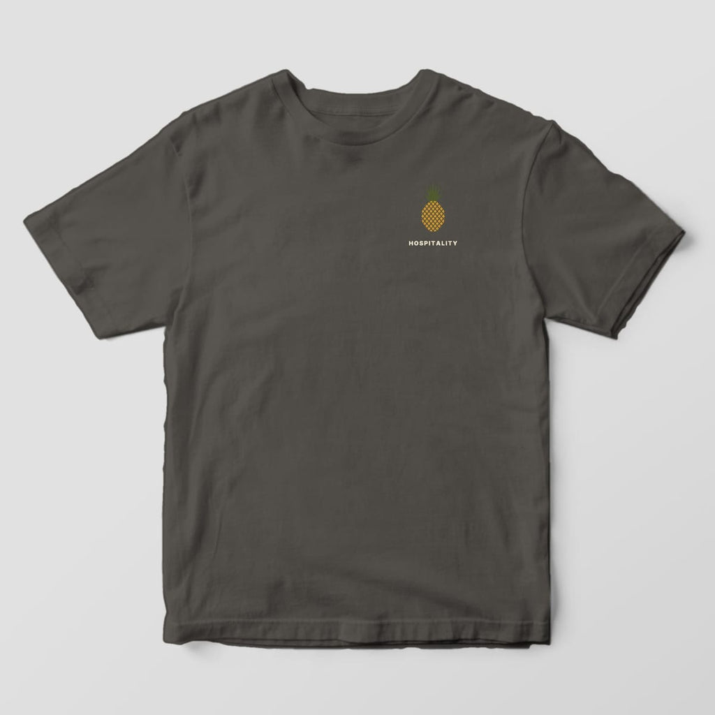 Front of a Pepper Comfort Colors 1717 tee with a small left-chest gold pineapple with an olive crown above the word HOSPITALITY.