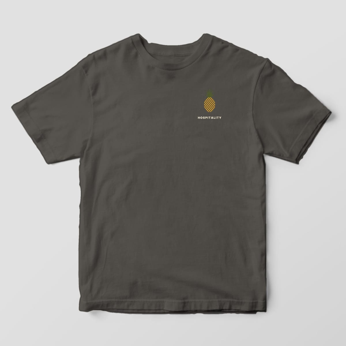Front of a Pepper Comfort Colors 1717 tee with a small left-chest gold pineapple with an olive crown above the word HOSPITALITY.