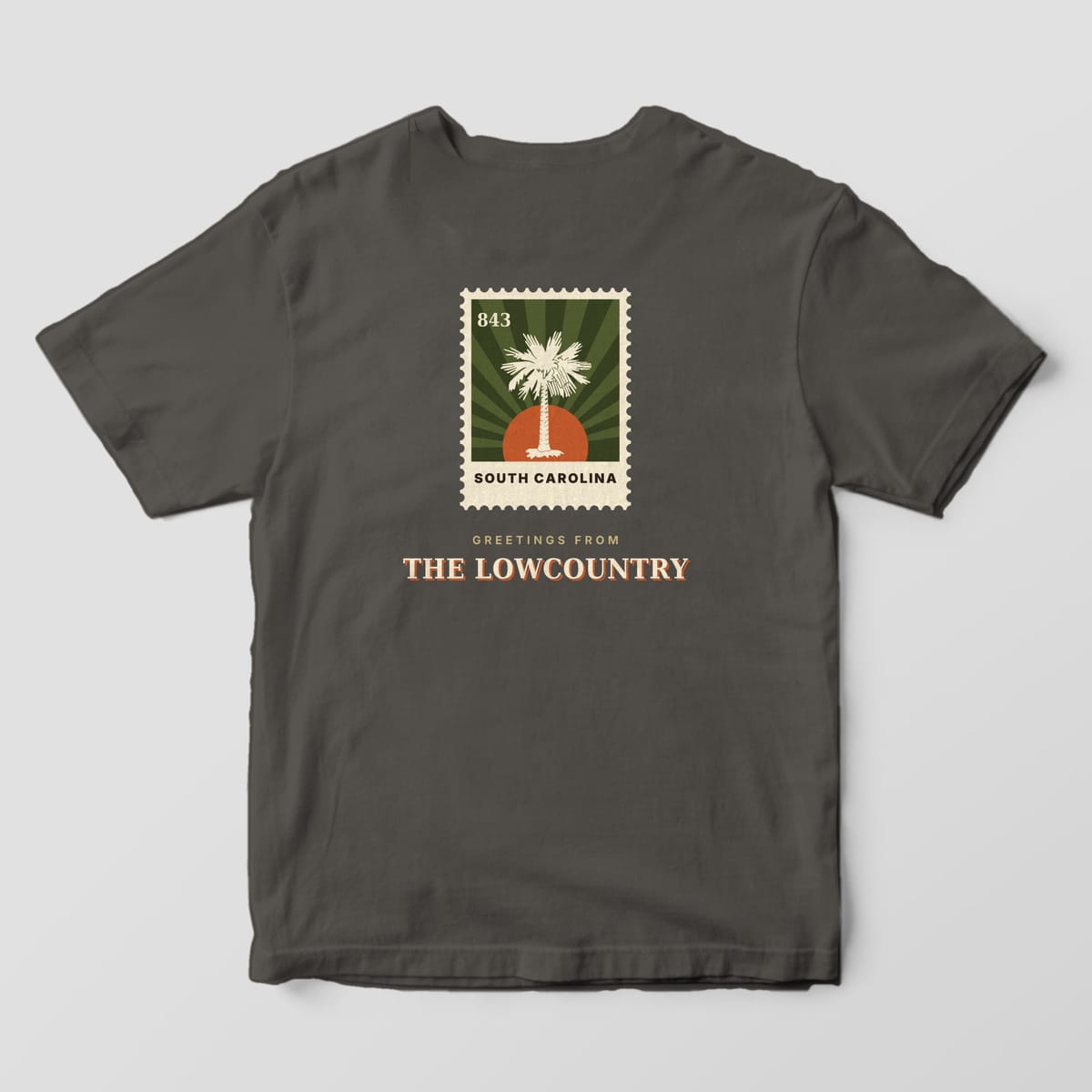 Back of a Pepper Comfort Colors 1717 tee with a perforated postage stamp showing a cream palmetto, orange sun and olive sunburst with 843 and SOUTH CAROLINA, above GREETINGS FROM THE LOWCOUNTRY.