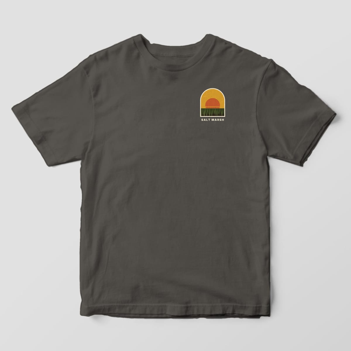 Front of a Pepper Comfort Colors 1717 tee with a small left-chest arch with a gold sky, orange sun and green marsh grass above the words SALT MARSH.