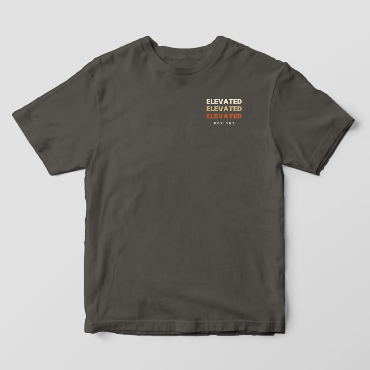 Front of a Pepper Comfort Colors 1717 tee with a small left-chest stack of ELEVATED three times in cream, tan and rust above DESIGNS.