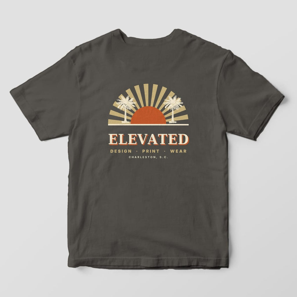Back of a Pepper Comfort Colors 1717 tee with a rust half-sun with tan sunburst rays and two cream palmetto trees, above ELEVATED, DESIGN · PRINT · WEAR and CHARLESTON, S.C.