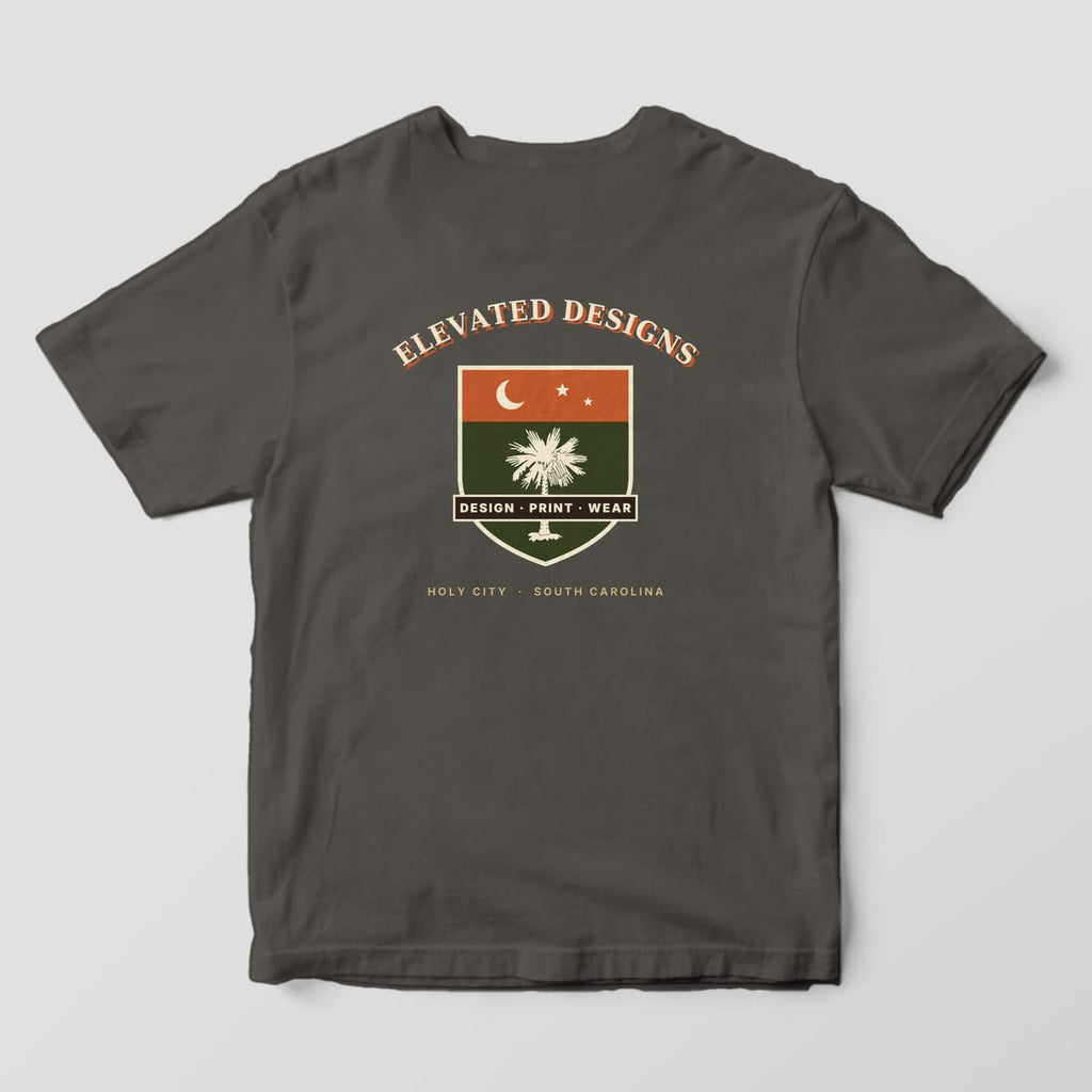 Back of a Pepper Comfort Colors 1717 tee with ELEVATED DESIGNS arched over a rust and olive shield with a crescent moon, stars, a cream palmetto and a DESIGN · PRINT · WEAR banner, above HOLY CITY · SOUTH CAROLINA.