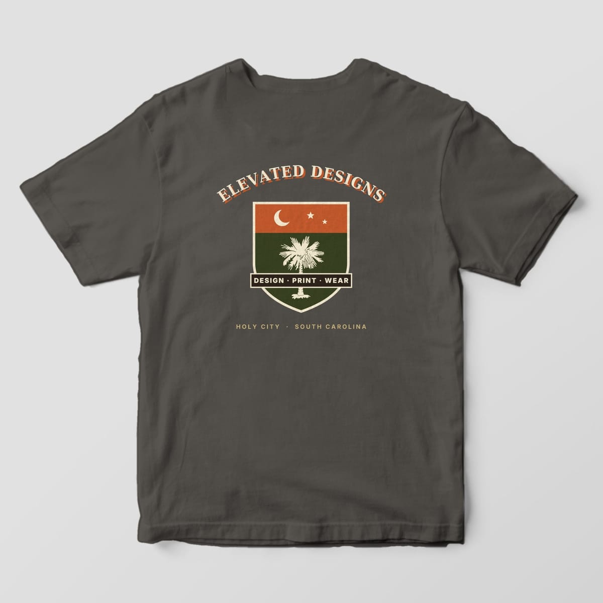 Back of a Pepper Comfort Colors 1717 tee with ELEVATED DESIGNS arched over a rust and olive shield with a crescent moon, stars, a cream palmetto and a DESIGN · PRINT · WEAR banner, above HOLY CITY · SOUTH CAROLINA.