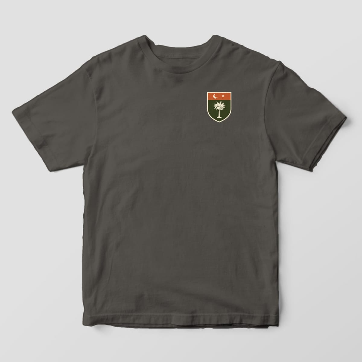 Front of a Pepper Comfort Colors 1717 tee with a small left-chest shield with a rust top band, cream crescent moon and star, and a cream palmetto on olive.